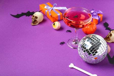 Glass of tasty cocktail with disco ball, gift boxes and Halloween decor on purple backgroundの写真素材