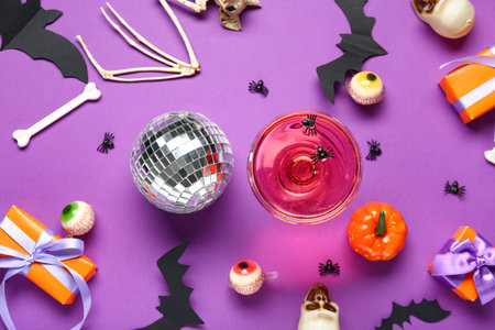 Glass of tasty cocktail with disco ball, gift boxes and Halloween decor on purple backgroundの写真素材