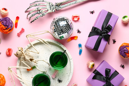 Glasses of tasty cocktails with disco ball, bat skeleton, cupcakes, gift boxes and Halloween decor on pink backgroundの写真素材