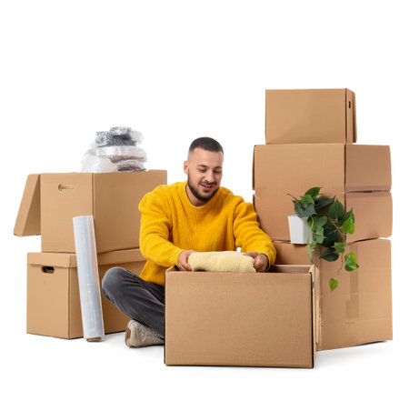 Young man unpacking moving box with clothes on white background