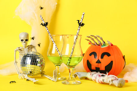 Glasses of tasty cocktails with disco ball, skeleton, bones and Halloween decor on yellow backgroundの写真素材