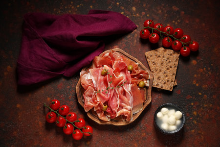 Wooden board with delicious prosciutto slices, olives, cherry tomatoes and bowl of mozzarella cheese on dark backgroundの写真素材