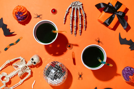 Plastic cups of tasty cocktails with disco ball, cupcakes, gift box, skeleton and Halloween decor on orange backgroundの写真素材