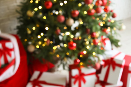 Blurred view of Christmas tree with glowing lights, baubles and gifts near white wallの写真素材