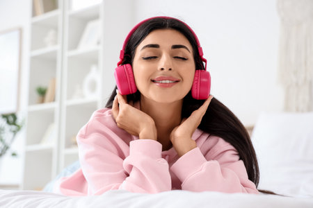 Beautiful young woman with modern headphones listening to music on bed in bedroomの写真素材