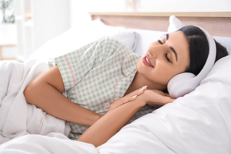 Beautiful young woman with modern headphones listening to music on bed in bedroomの写真素材