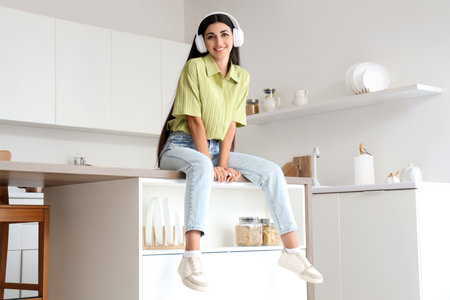 Beautiful young woman with modern headphones listening to music on table in kitchenの写真素材