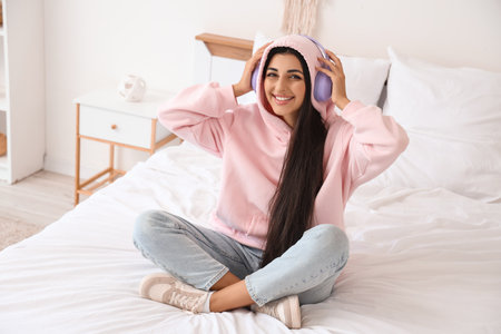 Beautiful young woman with modern headphones listening to music on bed in bedroomの写真素材