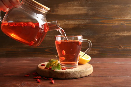 Pouring hot goji tea from teapot into glass cup on wooden backgroundの写真素材