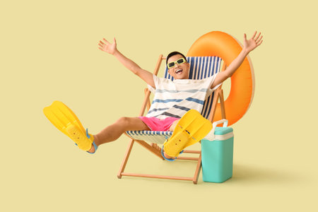 Young Asian man with beach cooler, flippers and inflatable ring sitting on deckchair against green backgroundの写真素材