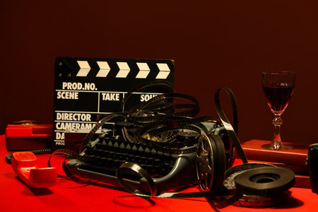 Vintage typewriter, telephone, film reel, clapperboard and glass of wine on red table near brown wall, closeupの写真素材