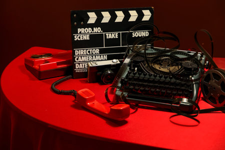 Vintage typewriter, telephone, film reel, clapperboard and photo camera on red table near brown wall, closeupの写真素材