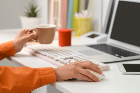 Hands of woman with computer mouse and cup of coffee at table in officeの写真素材