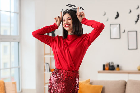 Beautiful young happy woman dressed for Halloween showing devil horns gesture in living roomの写真素材