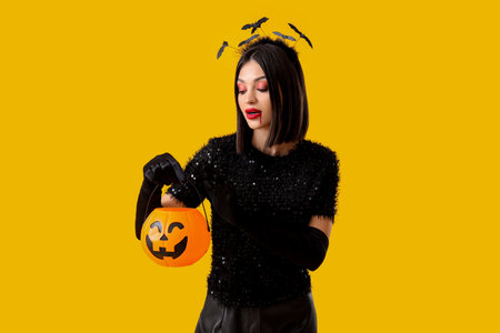 Beautiful young woman in bats headband dressed for Halloween with pumpkin on yellow backgroundの写真素材