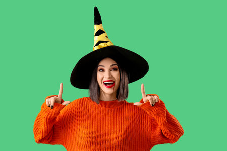 Beautiful young happy woman dressed as witch for Halloween pointing at something on green backgroundの写真素材