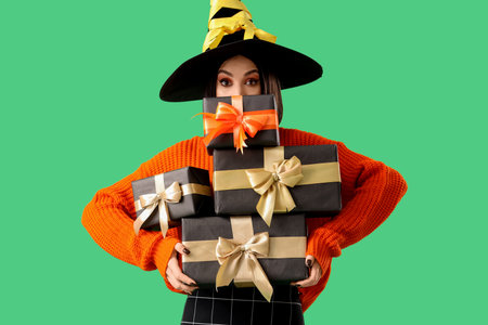 Beautiful young happy woman dressed as witch for Halloween with gift boxes on green backgroundの写真素材