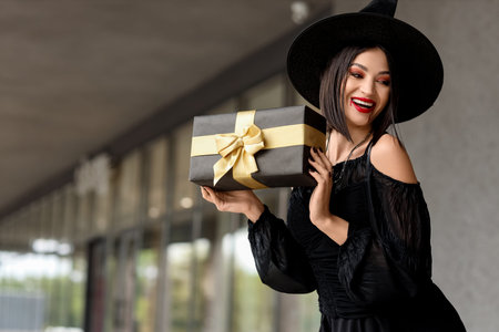 Beautiful young happy woman dressed as witch for Halloween with gift box on streetの写真素材