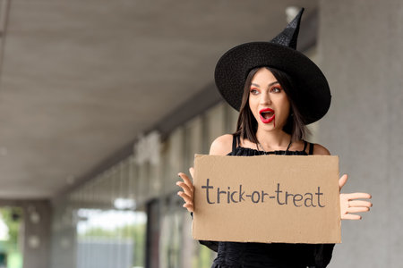 Beautiful young shocked woman dressed as witch for Halloween holding cardboard with text TRICK OR TREAT on streetの写真素材
