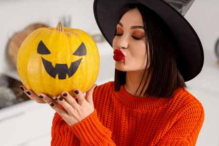 Beautiful young happy woman dressed as witch for Halloween with pumpkin blowing kiss in kitchenの写真素材