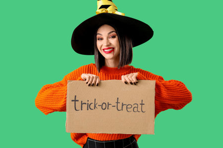 Beautiful young happy woman dressed as witch for Halloween holding cardboard with text TRICK OR TREAT on green backgroundの写真素材