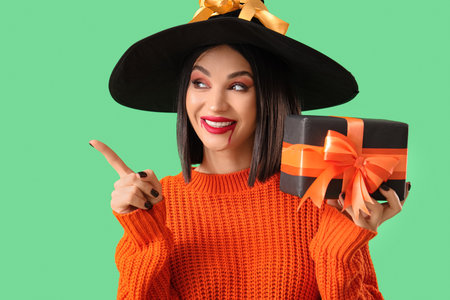 Beautiful young happy woman dressed as witch for Halloween with gift box pointing at something on green backgroundの写真素材