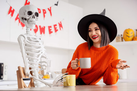 Beautiful young happy woman dressed as witch for Halloween drinking tea with skeleton at table in kitchenの写真素材