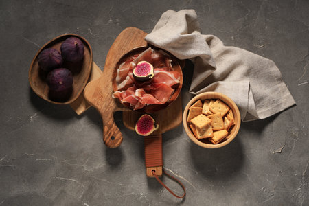 Wooden plate with delicious prosciutto slices, fresh fig and bowl of crackers on gray backgroundの写真素材