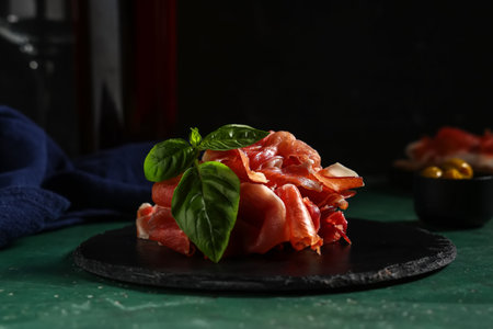 Black board with delicious prosciutto slices and basil leaves on green tableの写真素材