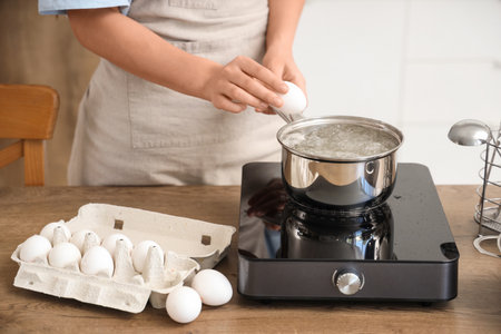 Woman putting egg into saucepan with boiling water in kitchenの写真素材