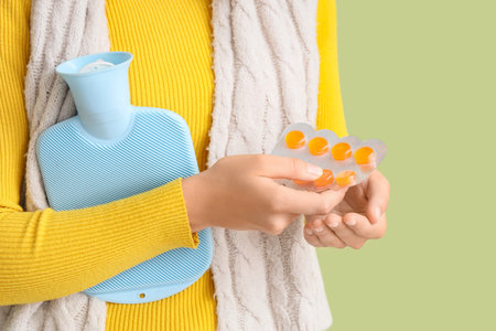Ill young woman with hot water bottle taking throat lozenges on green background, closeupの写真素材