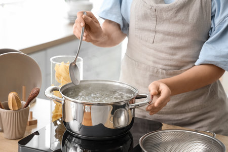 Woman and stainless steel pot with boiling water on stove in kitchen, closeupの写真素材
