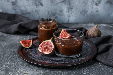 Plate with glass and jar of sweet fig jam on gray grunge background