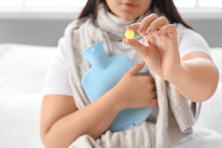 Ill young woman with throat lozenge and hot water bottle in bedroom, closeupの写真素材