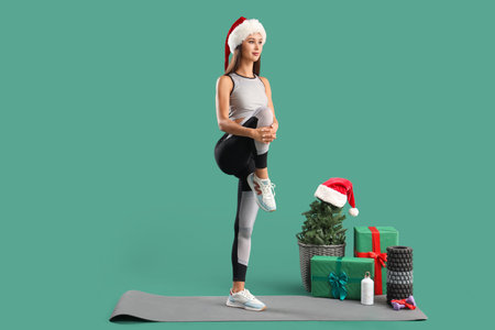 Sporty young woman in Santa hat with Christmas presents training on green backgroundの写真素材