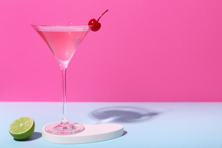 Glass of tasty Cosmopolitan cocktail garnished with cherry and lime on blue table against pink backgroundの写真素材