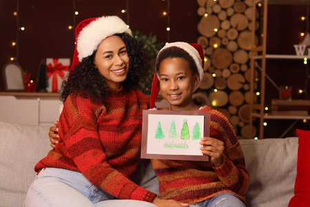 African-American mother and her daughter in Santa hats with greeting card at home on Christmas Eveの写真素材