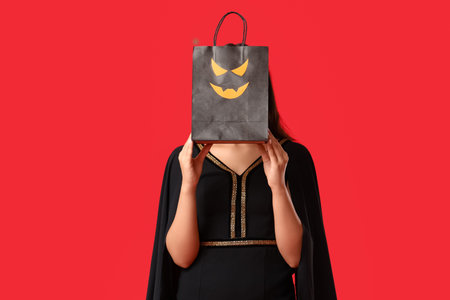 Beautiful young woman dressed as witch for Halloween with shopping bag of candy corns on red backgroundの写真素材