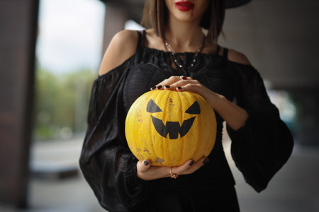 Beautiful young woman dressed as witch for Halloween with pumpkin on street, closeupの写真素材