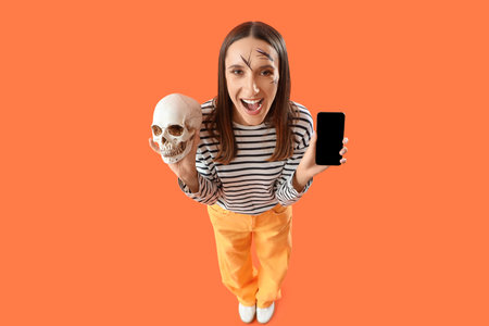 Young woman with Halloween human skull and mobile phone on orange background