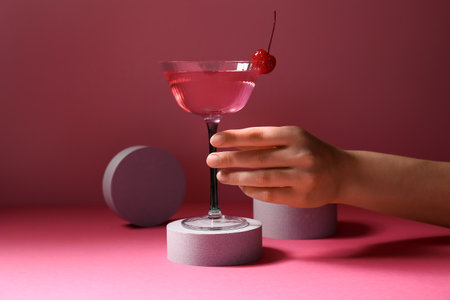 Female hand with glass of tasty Cosmopolitan cocktail and plaster podiums on pink backgroundの写真素材