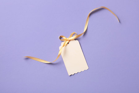 Blank tag with bow made of golden ribbon on purple backgroundの写真素材
