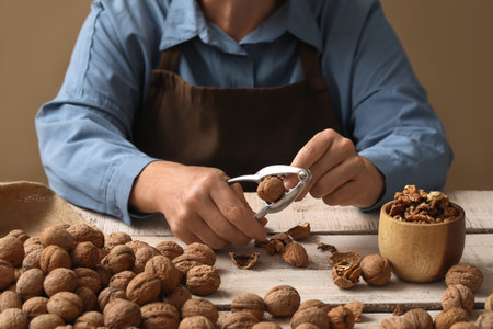 Elderly woman cracking walnuts with nutcracker at wooden table against beige wallの写真素材