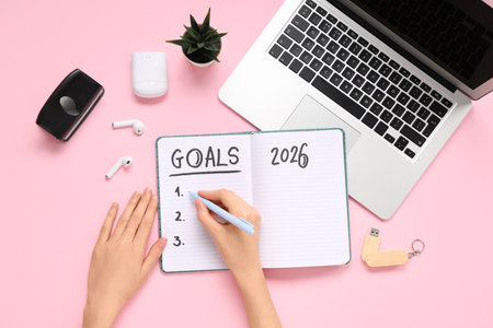 Female hands writing 2026 goals in notebook with laptop on pink backgroundの写真素材