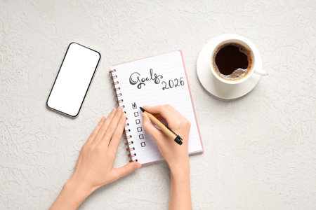Female hands writing 2026 goals in notebook with mobile phone and coffee on white grunge backgroundの写真素材