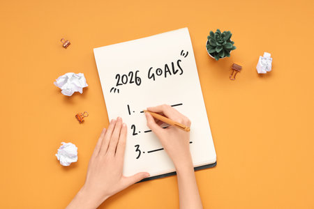 Female hands writing 2026 goals in notebook with crumpled paper on orange backgroundの写真素材