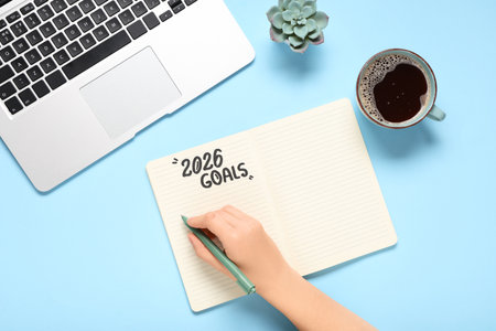 Female hand writing 2026 goals with coffee cup and laptop on blue backgroundの写真素材