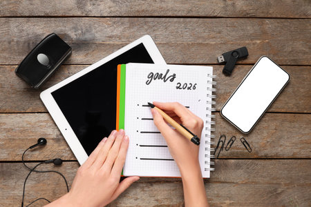 Female hands writing 2026 goals in notebook with gadgets on wooden background