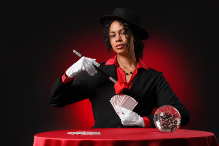 Young African-American magician with magic wand, cards and crystal ball on table against dark red backgroundの写真素材