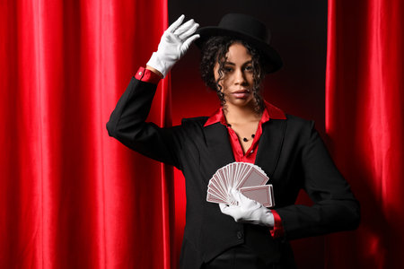 Young African-American magician with cards near red curtains on dark backgroundの写真素材